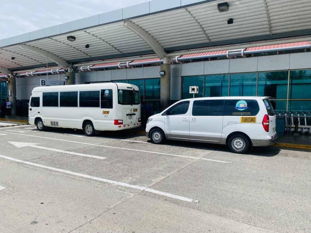 Two white private shuttle vans parked at SJO airport terminal, ready for guest pickups in Costa Rica